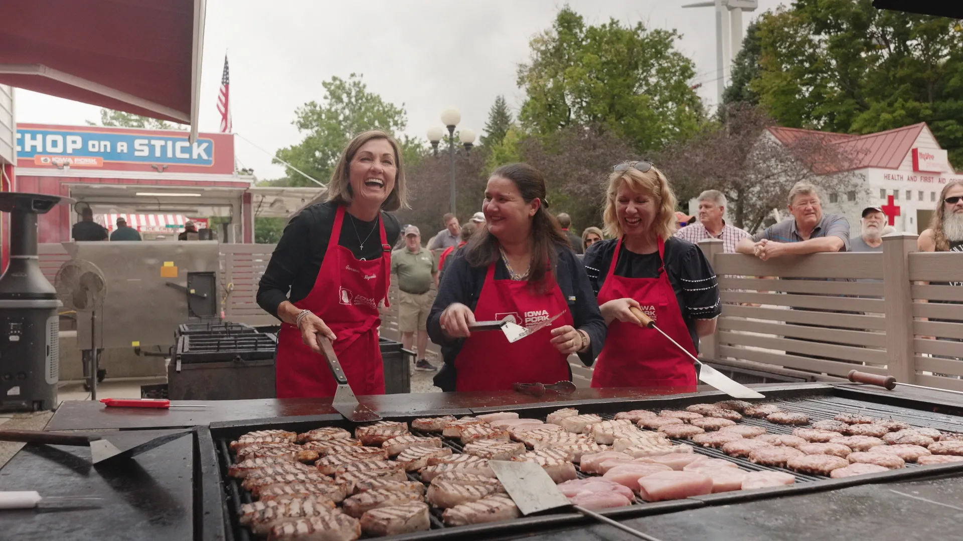 Three people in red aprons standing in front of a huge grill covered in sausages