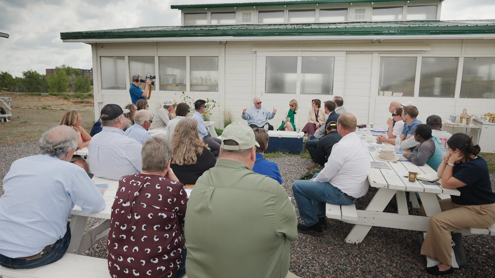 Group of people sitting outside in a circle of picnic tables with one man speaking with his arms raised